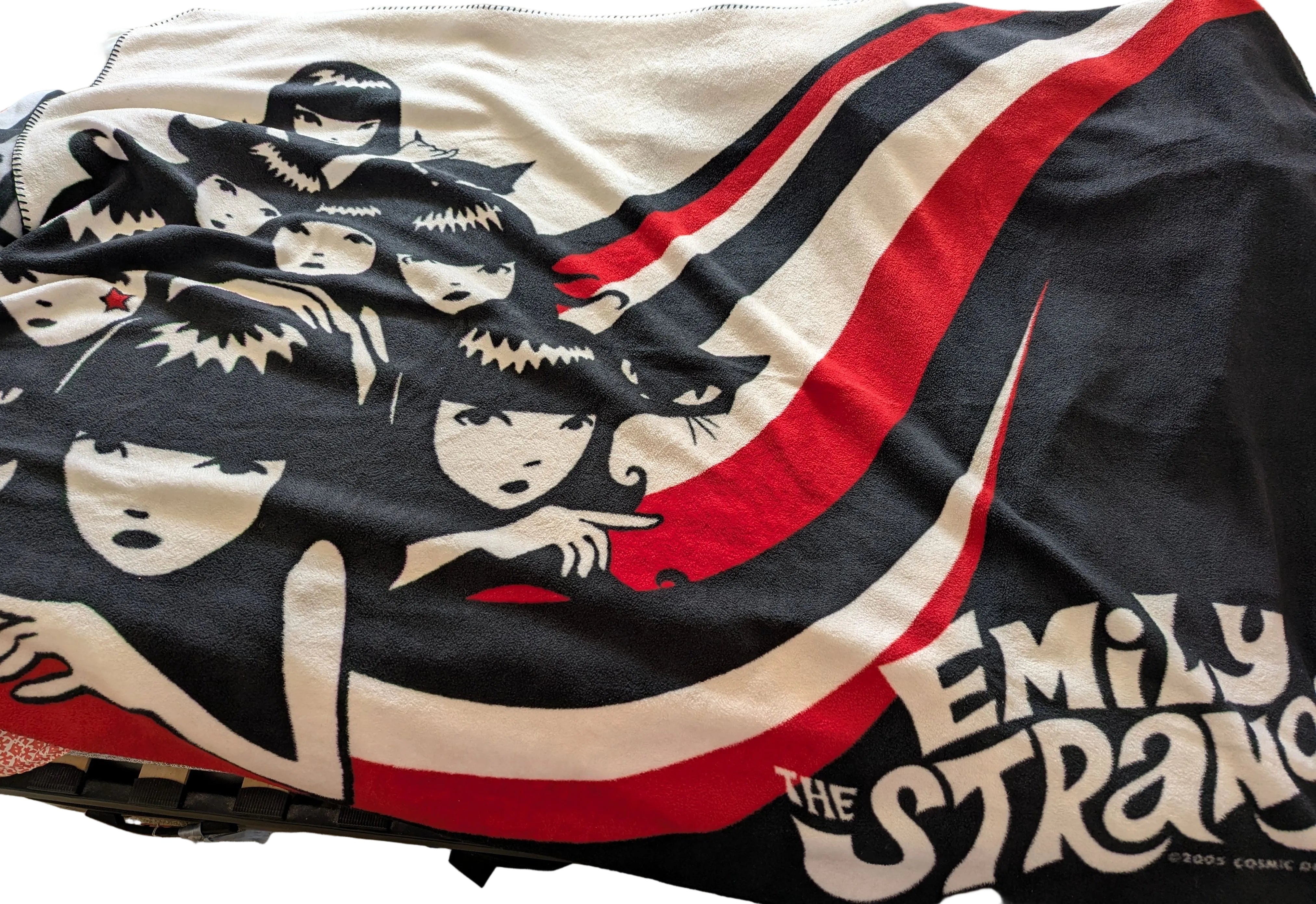 A close-up of a printed fabric item, likely a blanket or towel, featuring stylized anime characters and the text 'EMILY THE STRANGE'. The design uses black, off-white, and red