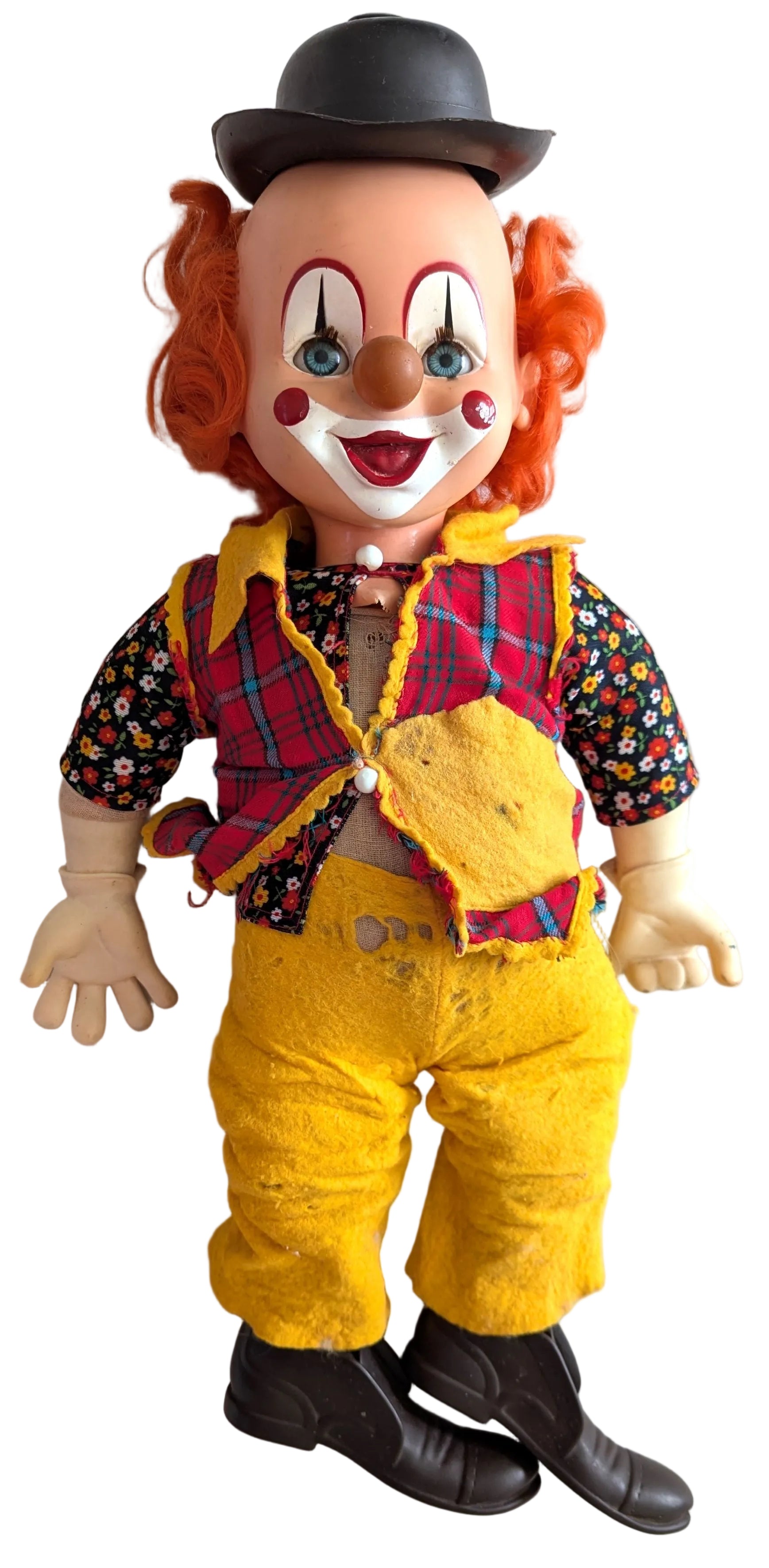 A vintage clown doll with orange curly hair, a black bowler hat, a red and yellow plaid vest, a floral short-sleeve shirt, yellow pants, and black shoes, photographed against a white paneled wall.
