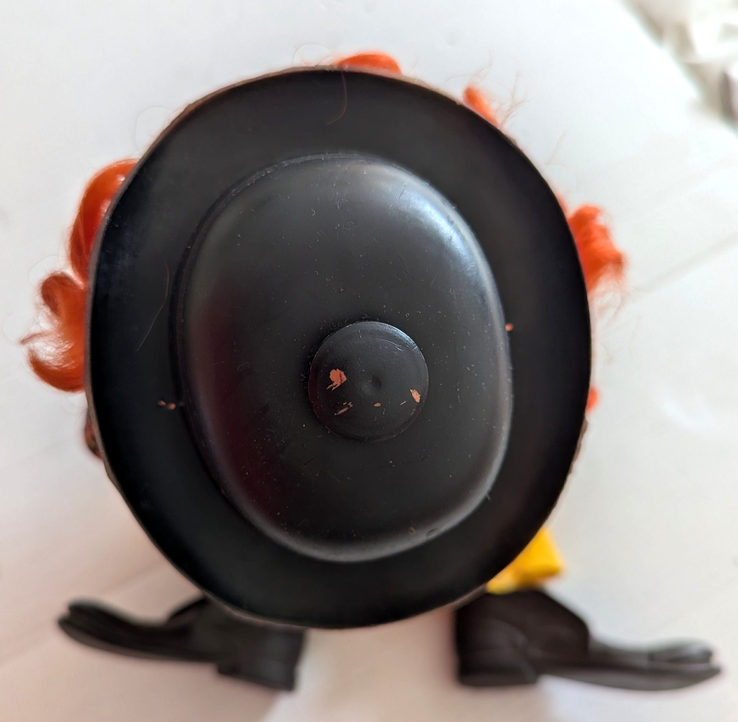 A close-up top-down view of a toy or doll's head and hat, featuring a large black hat, orange hair, and black shoes on a white background.