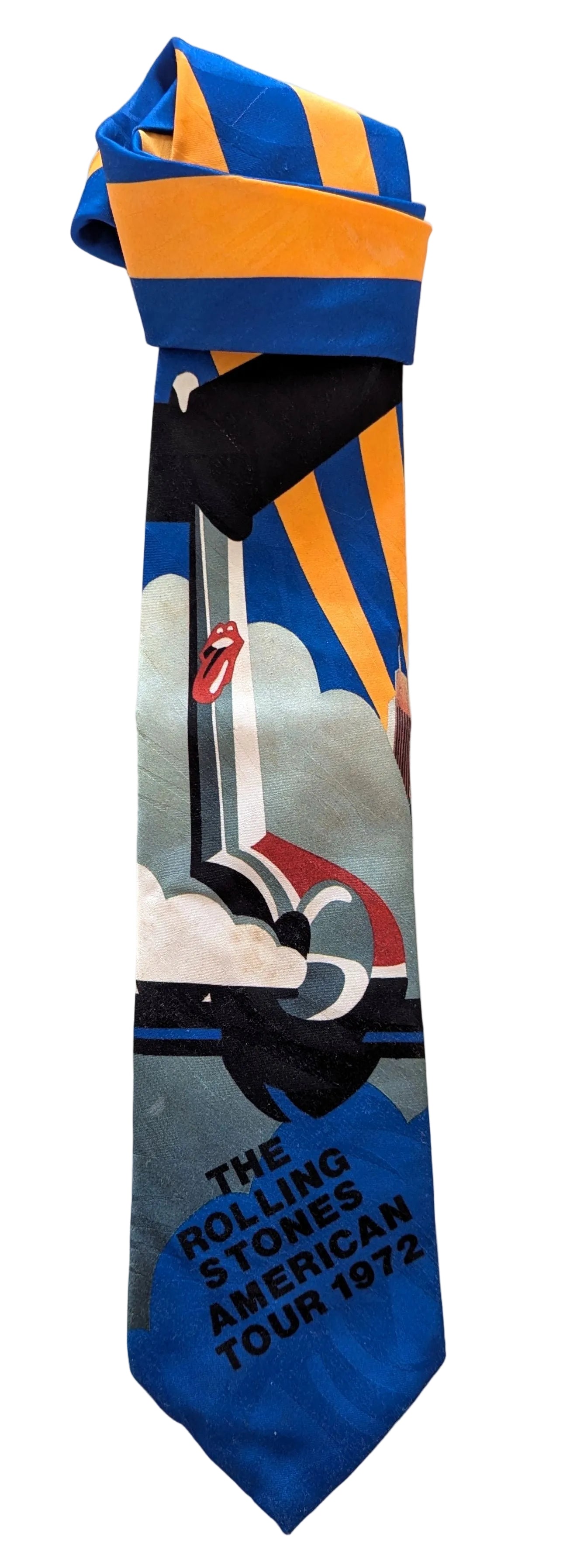 A rolled-up necktie featuring a graphic print of the Rolling Stones 'American Tour 1972' with blue, orange, black, white, and gray elements, displayed on a light fabric background.