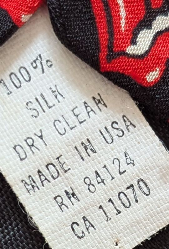 Close-up of a fabric care label reading '100% SILK DRY CLEAN MADE IN USA RN 84124 CA 11070' attached to a black and red patterned tie.