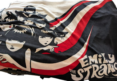 A close-up of a printed fabric item, likely a blanket or towel, featuring stylized anime characters and the text 'EMILY THE STRANGE'. The design uses black, off-white, and red