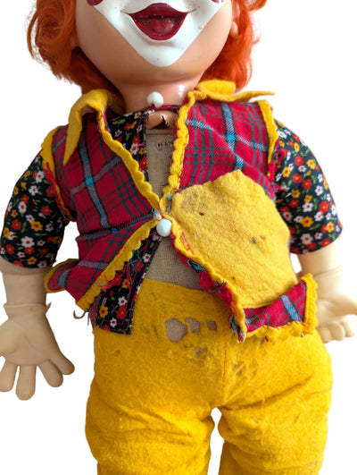 A vintage-style doll or collectible figure with orange hair, a white face, and a handmade patchwork outfit featuring a red plaid vest, floral short-sleeve shirt, and yellow pants, photographed against a white background.