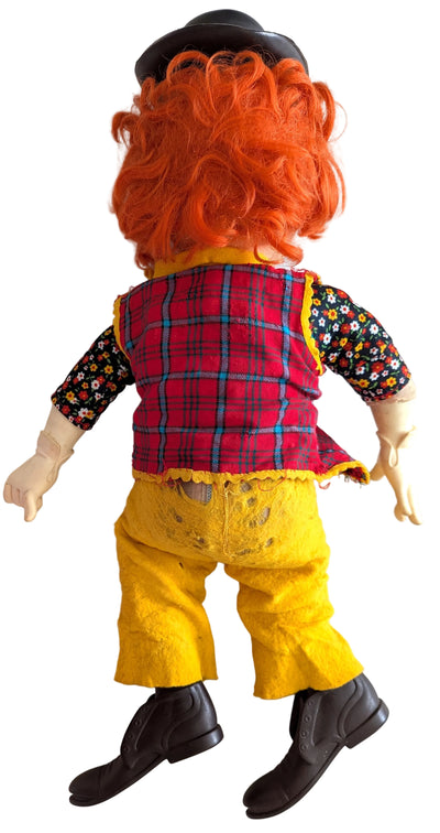 A vintage doll viewed from the back, featuring bright orange curly hair, a black hat, a red and blue plaid vest, a black floral long-sleeve shirt, yellow pants, and black boots, lying on a white background.