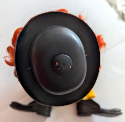 A close-up top-down view of a toy or doll's head and hat, featuring a large black hat, orange hair, and black shoes on a white background.