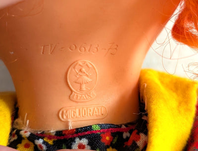 Close-up of a vintage Italian doll's neck and upper torso, showing the molded 'MIGLIORATI' logo and 'TV-0613-73' markings on the peach-colored plastic skin, wearing a yellow garment and a floral patterned skirt.