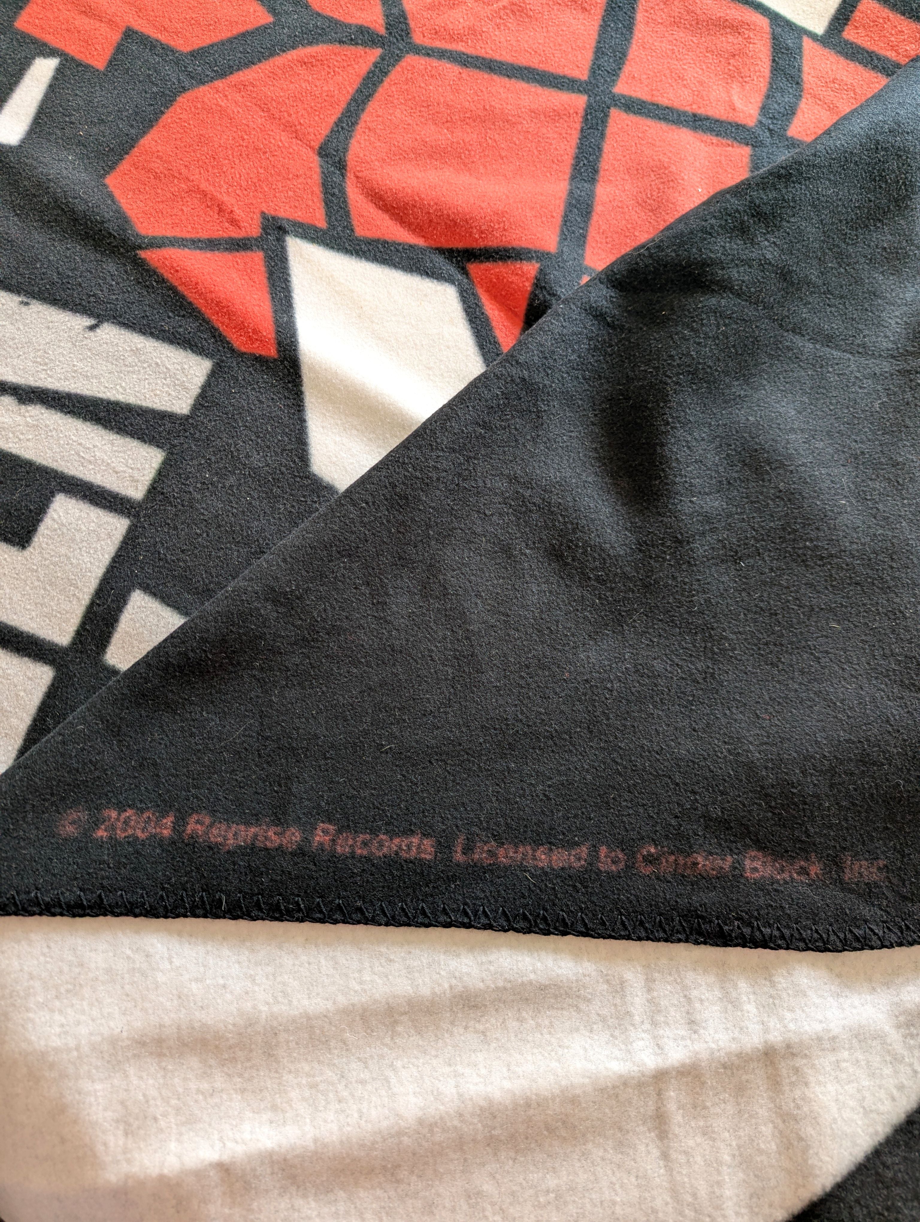 Close-up of a black fleece blanket with a red, white, and black geometric Cinder Block graphic and copyright text reading '2004 Reprise Records. Licensed to Cinder Block, Inc.