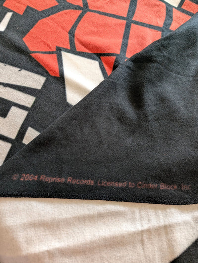 Close-up of a black fleece blanket with a red, white, and black geometric Cinder Block graphic and copyright text reading '2004 Reprise Records. Licensed to Cinder Block, Inc.
