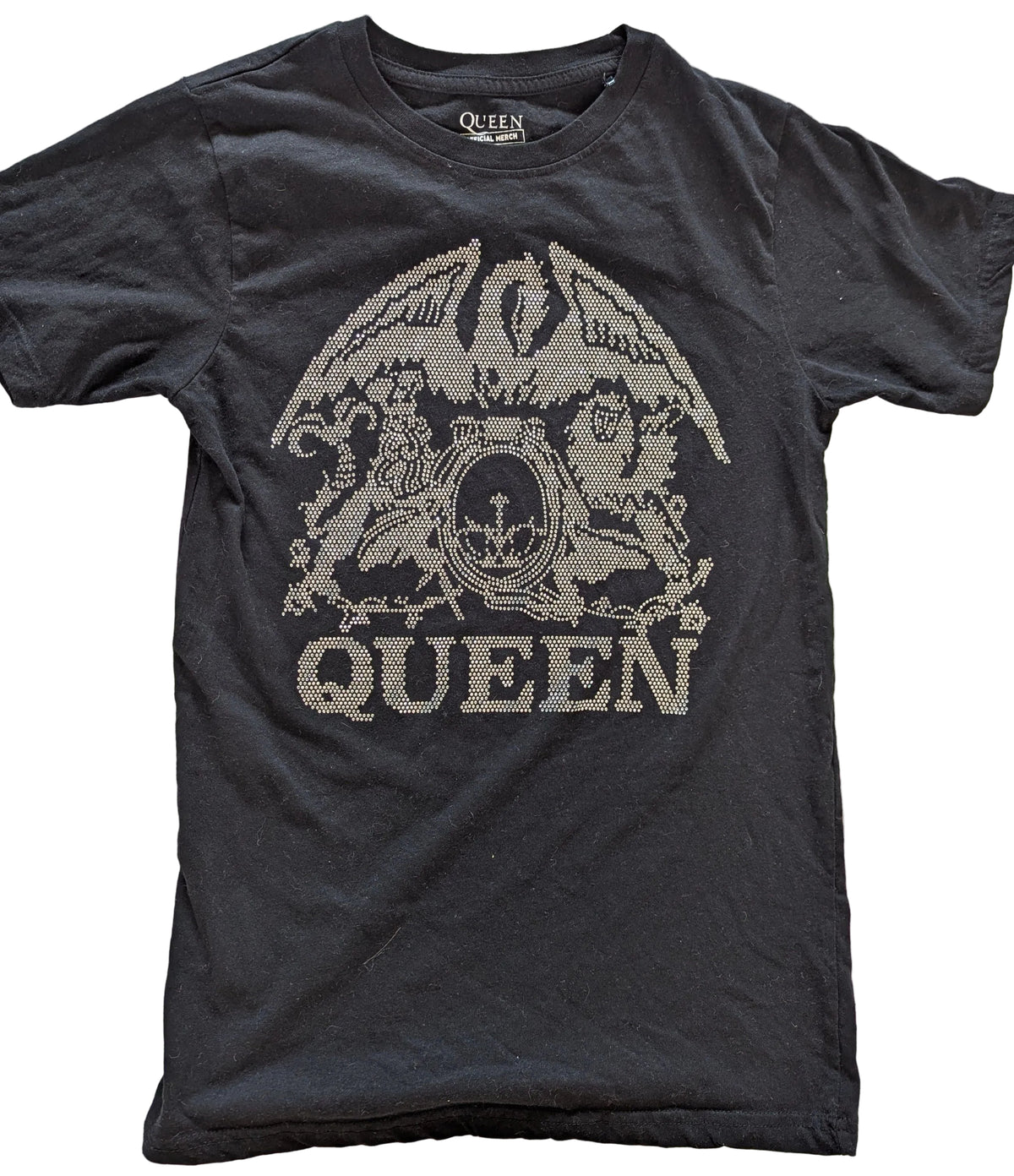 A black short-sleeve Queen band t-shirt featuring the band's logo and 'QUEEN' text in a distressed, pixelated silver graphic on the front, laid flat on a light wood background.