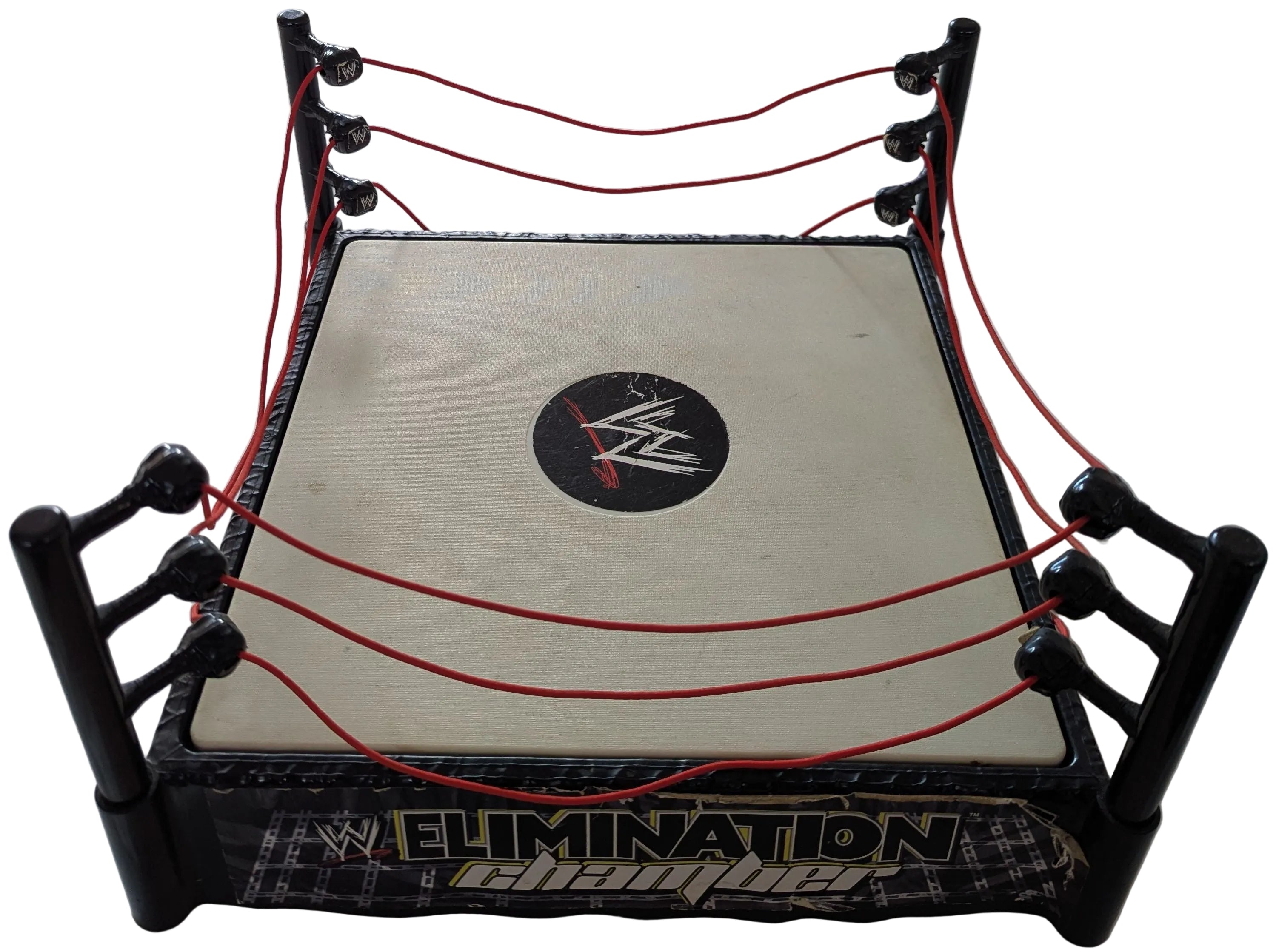 A miniature wrestling ring set on a white fabric surface, featuring black corner posts, red ropes, a cream-colored canvas top, and a central WWE Elimination Championship logo.